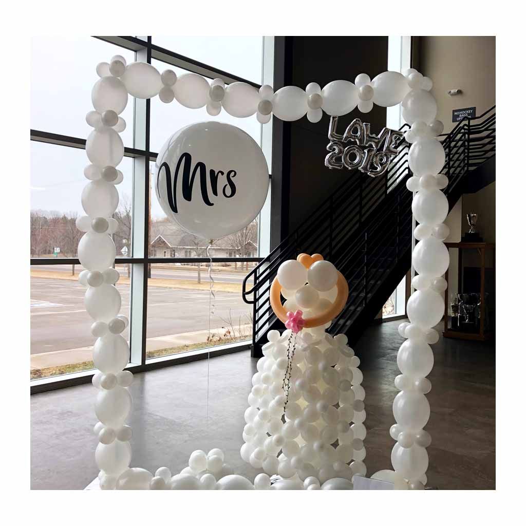 A decorative setup features a large white balloon arch framing a white balloon display resembling a wedding dress, accompanied by a balloon with "Mrs" written on it and a "Love" sign, set against a backdrop of large industrial-style windows.
