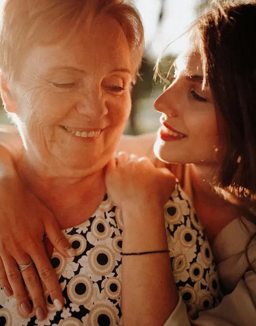 Older woman and younger woman sharing a warm, close moment together, both smiling in soft natural light.