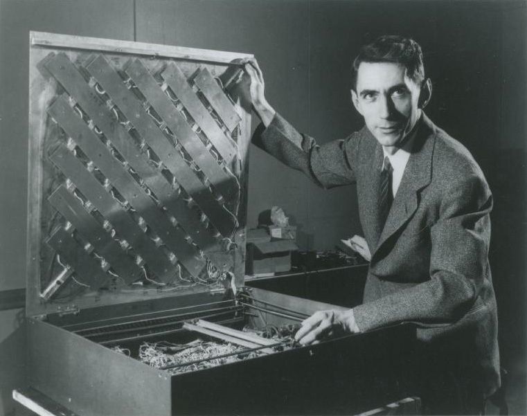 Black-and-white portrait of a suited man presenting an open experimental electronic device.