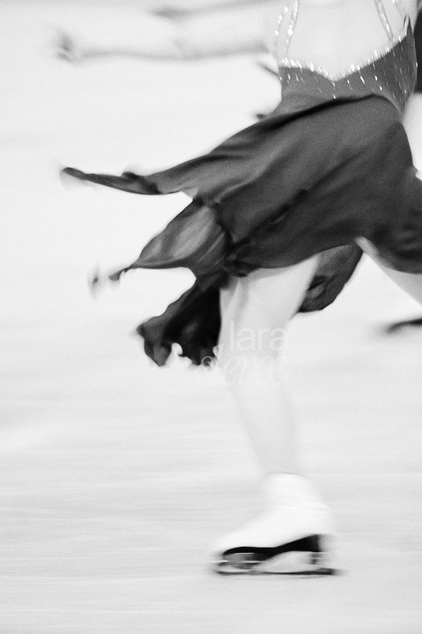 Schwarz-Weiß Bild im Analog-Stil einer Eiskusntläuferin. Ihr Rock durch den Fahrtwind verweht. Nur ihr Bein und der Rücken ist zu erkennen. Team Skyliners. ISU Synchronized Skating Championships 2026 in Salzburg. Fotografiert von Lara Slevec.