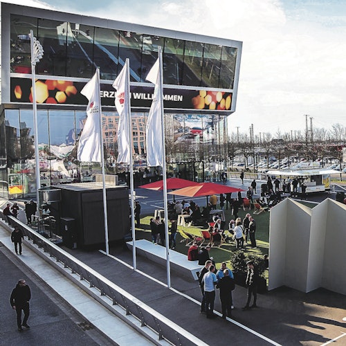 戶外場景，有人圍著座位在現代建築旁邊的傘下閒聊，建築物上有反光玻璃和國旗。