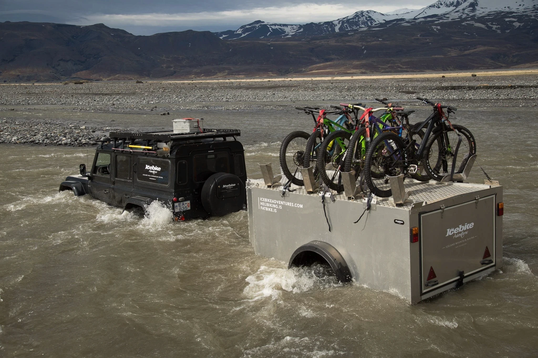 4x4 towing a bike trailer through a glacial river with mountains behind.