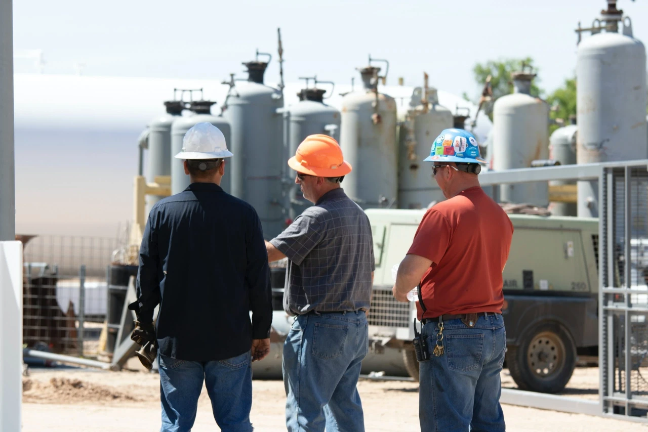 Three workers wearing hard hats stand in front of industrial equipment, with one pointing towards the machinery.