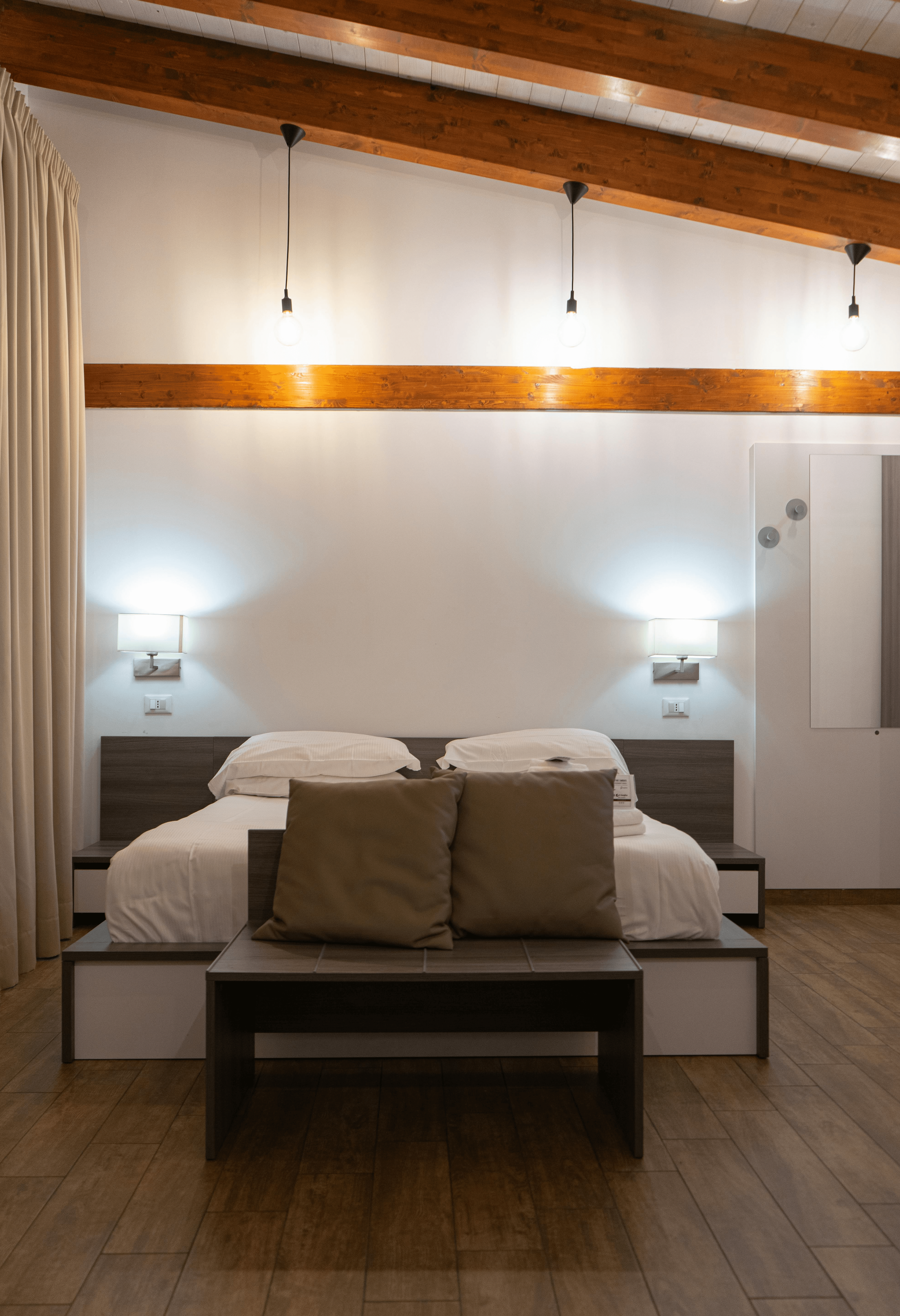 Imagem ccom uma cama e espelho de ferro em lados opostos, com  tons pasteis claros e traveseiros em cima da cama, o chão de madeira e uma grande janela com cortinhas finas, deixando o sol entrar