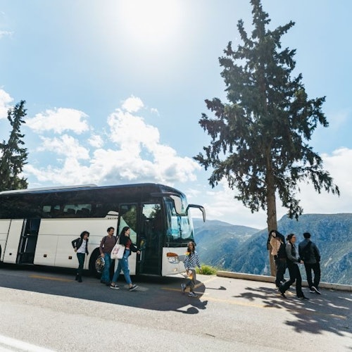 Pessoas caminhando ao lado de um ônibus de turismo estacionado em uma estrada de montanha cênica, com árvores altas e um céu limpo ao fundo.