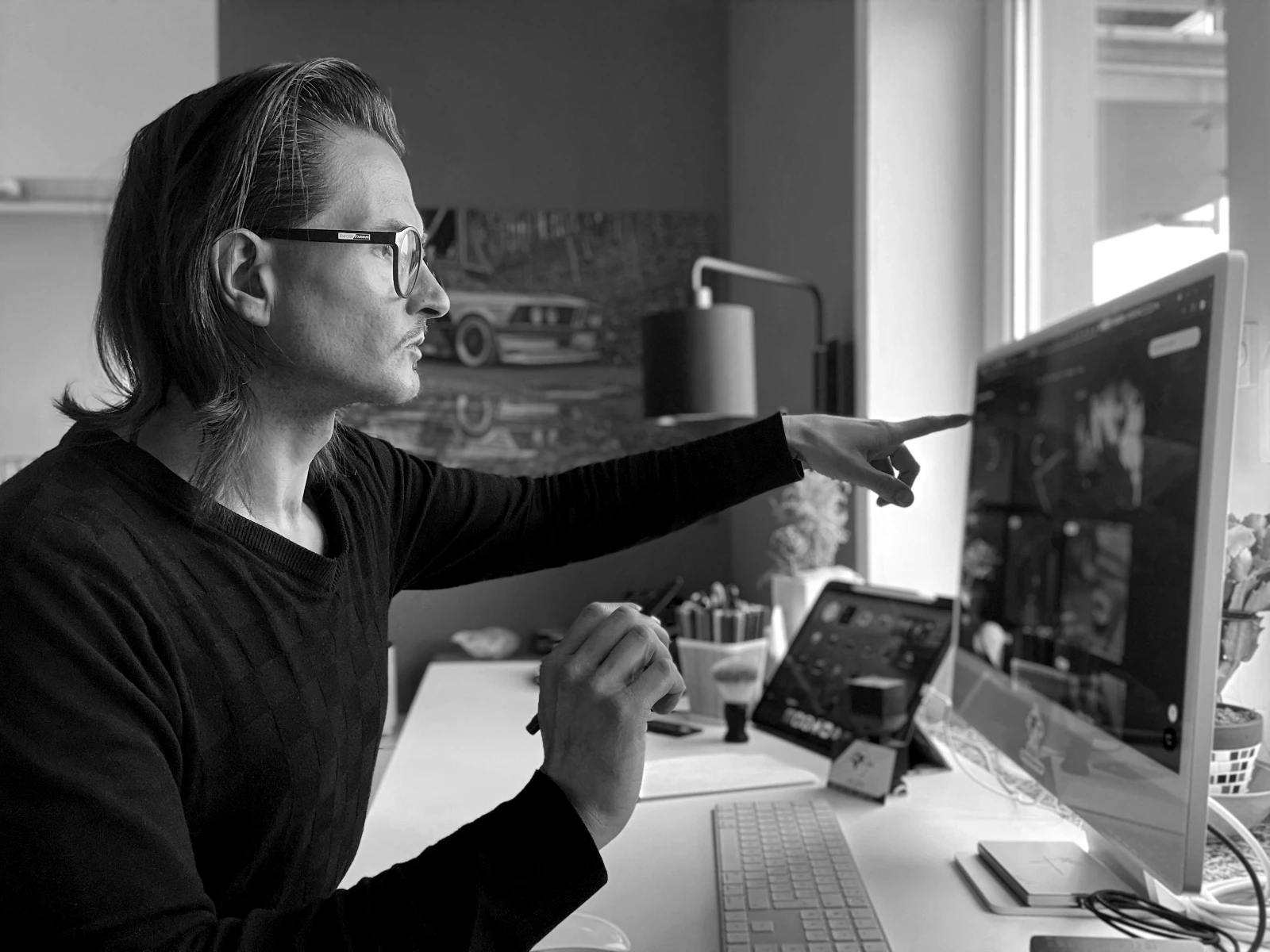 Jens Pfeifer arbeitet fokussiert an einem Apple iMac und zeigt mit dem Finger auf ein Interface-Detail auf dem Bildschirm. Seitliche Perspektive im Studio-Workspace mit iPad, Tastatur, Skizzenblock und Farbstiften auf dem Schreibtisch – visuelle Darstellung eines analytischen Design- und Review-Prozesses in einem professionellen, kreativen Arbeitsumfeld.