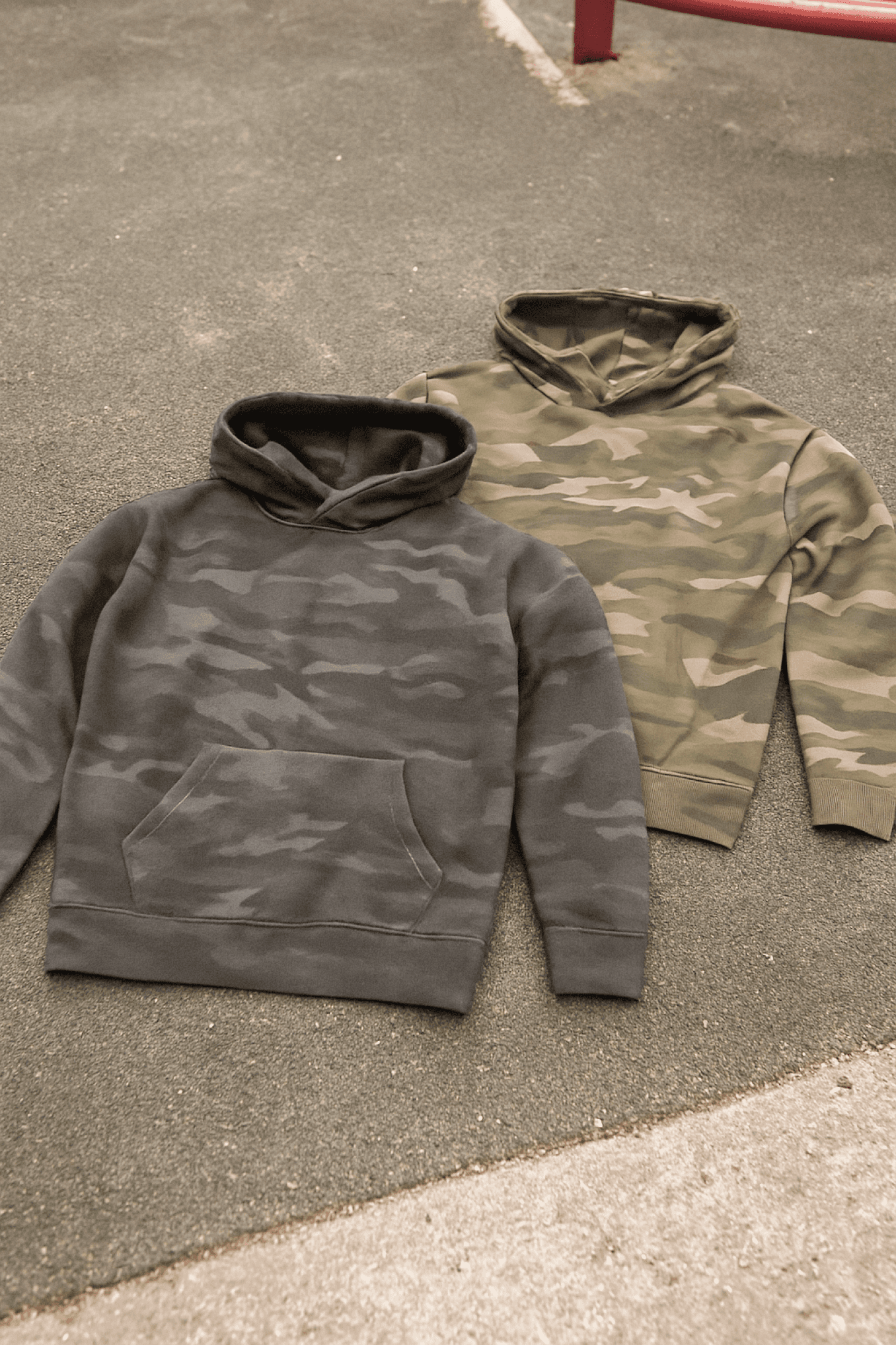 Two camouflage hoodies laid flat on a gray surface. One is darker and the other is lighter in color.