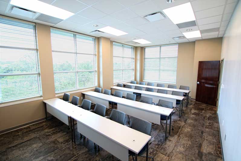 A spacious classroom with rows of tables, chairs, and large windows letting in natural light.