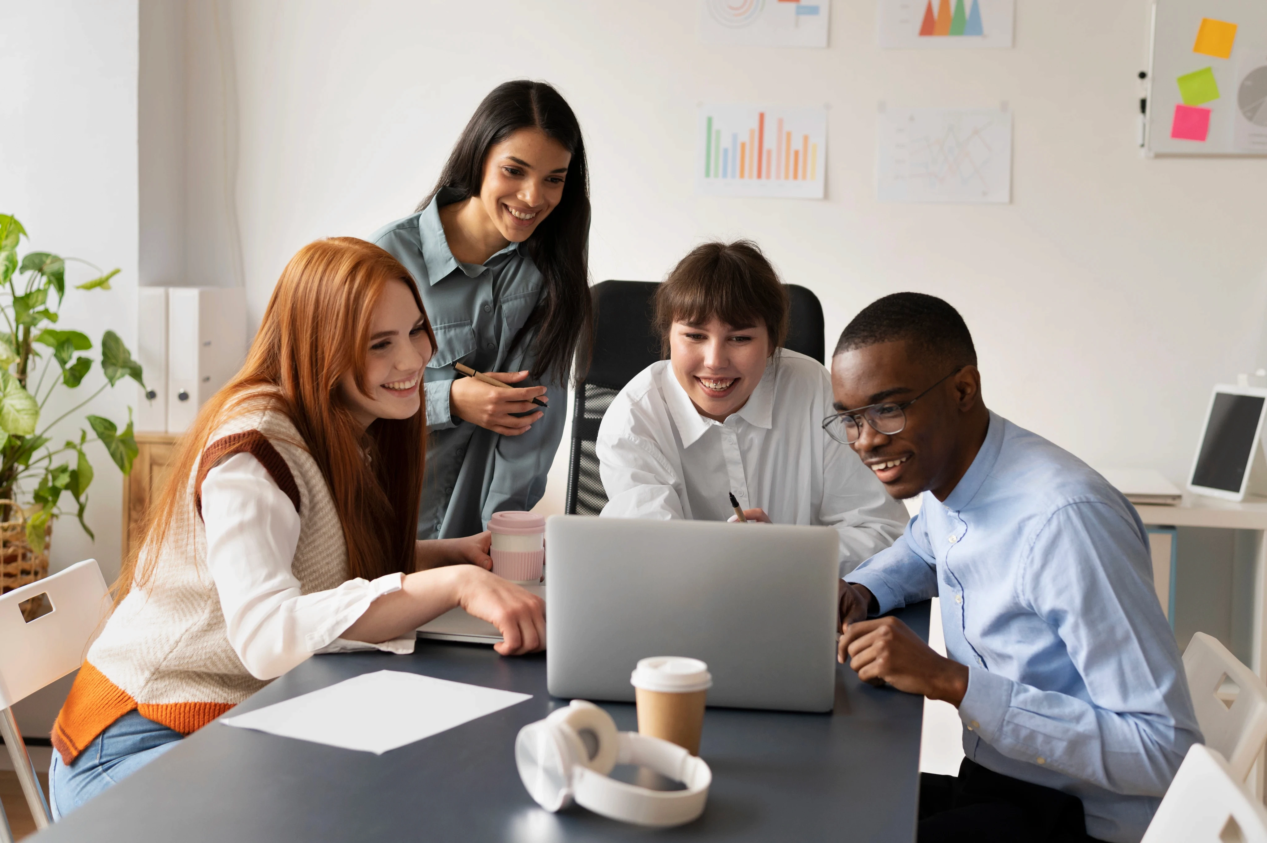 A diverse group of smiling coworkers collaborating and looking at a laptop screen together in a bright, modern office with data charts on the wall.