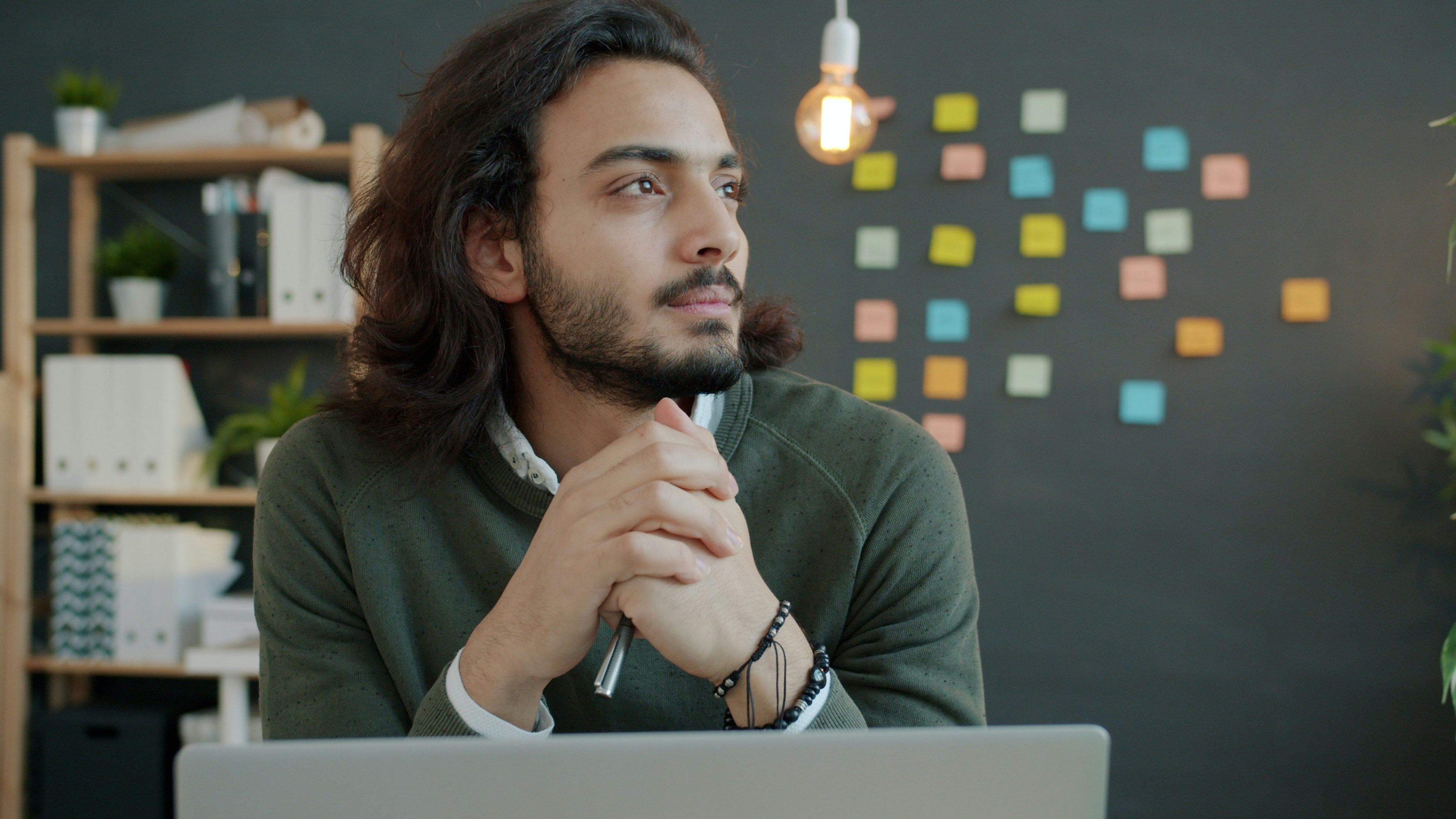 Man with long hair thinking at desk with laptop.