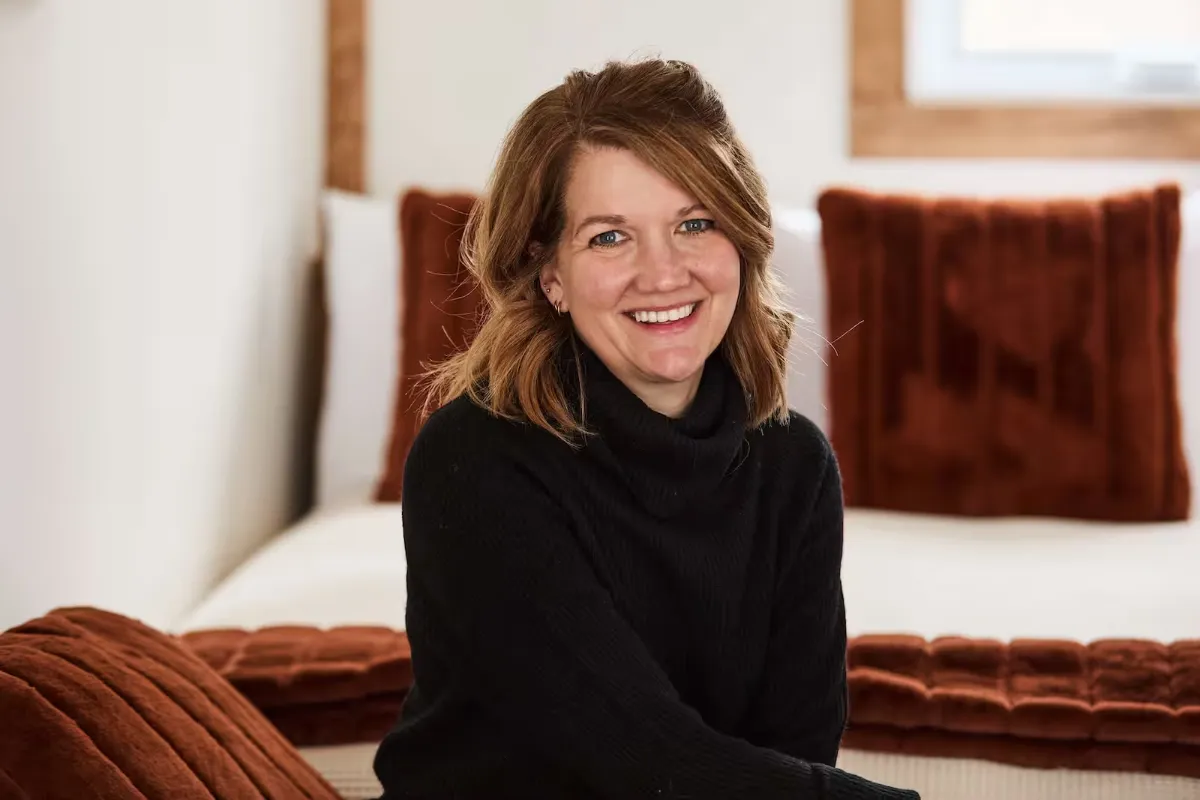 Kerri Gibson smiling with arms crossed, in a peach blazer against a wood wall