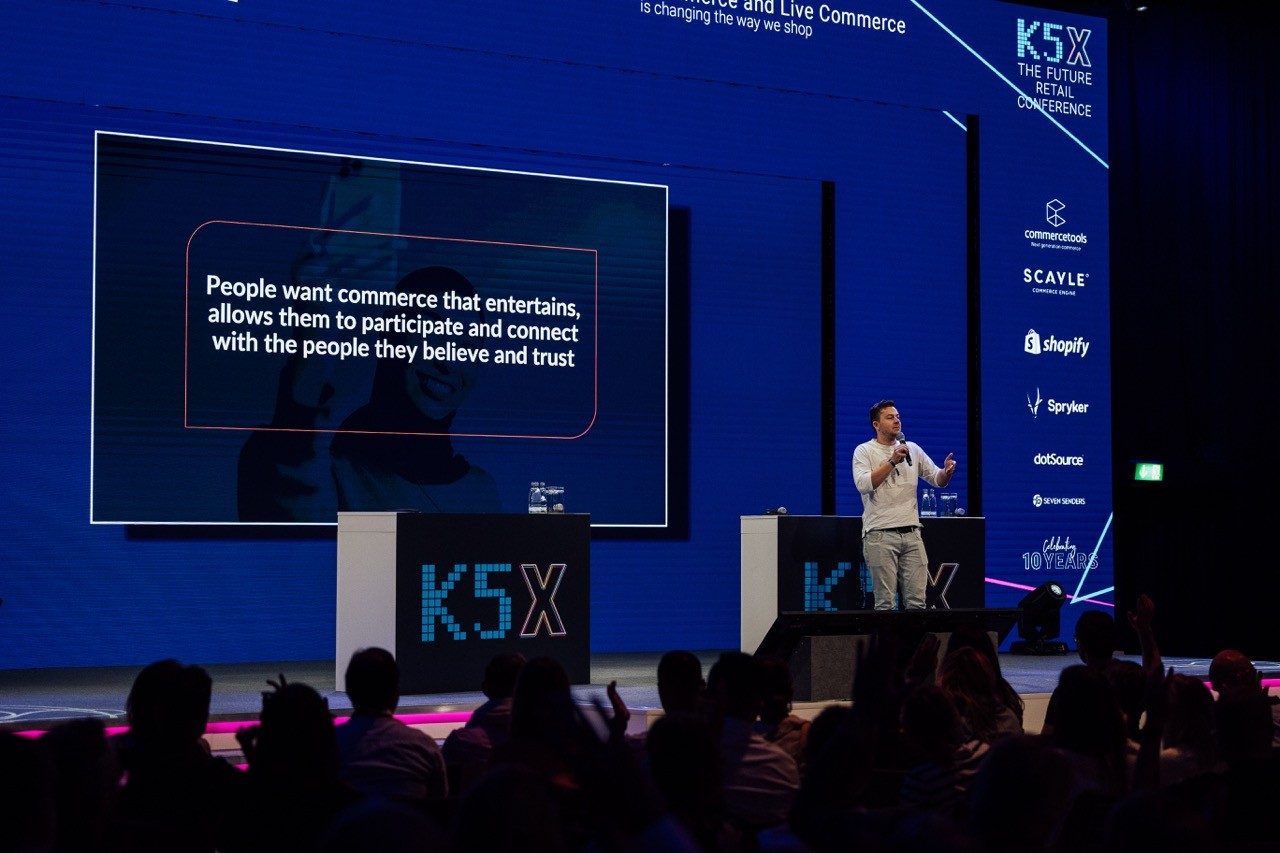 A person wearing glasses and a dark sweater sits at a desk, smiling and giving a thumbs up, with a laptop open in front of them, and a framed abstract painting hanging on the wall behind.