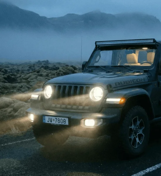 A dark Jeep Wrangler driving on an asphalt road in the fog at twilight, with headlights and roof lights on.
