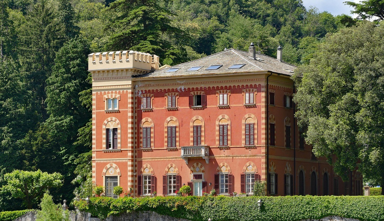 A large, red mansion with elegant brickwork and arched windows sits amid lush greenery. A turret on the left adds a castle-like feel. Serene and grand.
