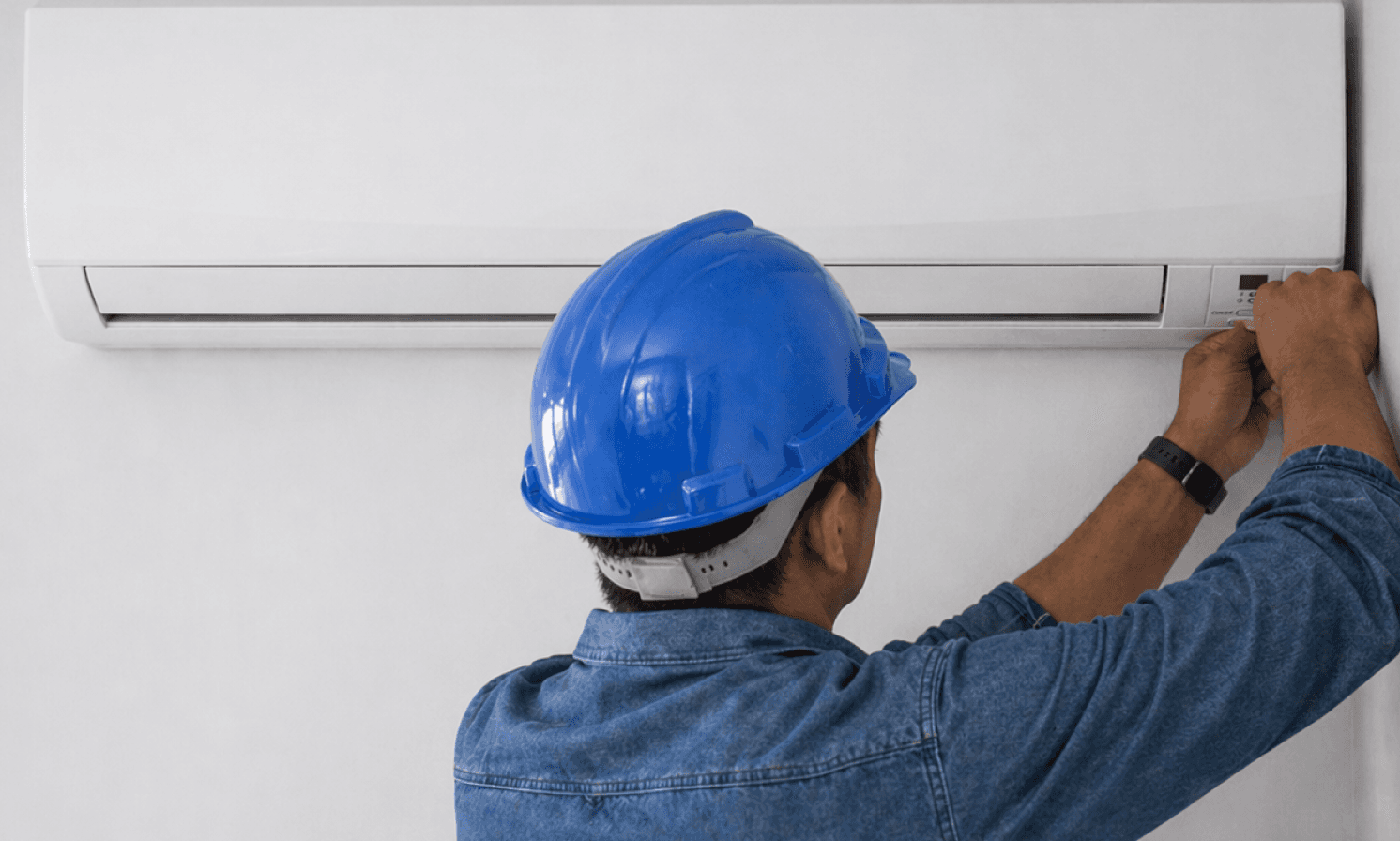 A technician in a blue hard hat adjusting a wall-mounted air conditioner.