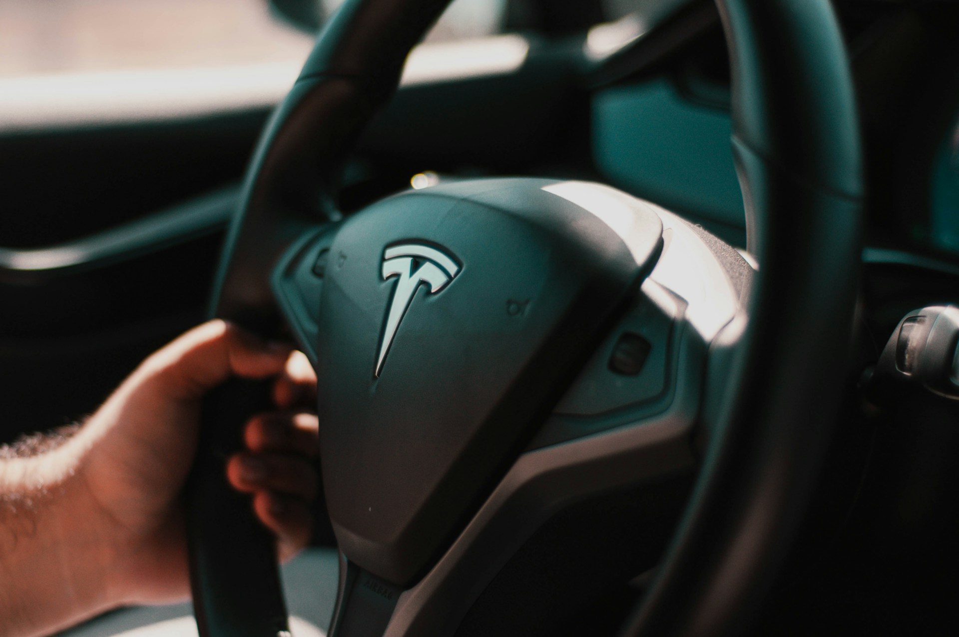  A close-up shot of the steering wheel of a Tesla car, clearly showing the metallic Tesla logo in the center. A person's hand is gripping the dark steering wheel on the left side. The lighting is bright, highlighting the interior details.