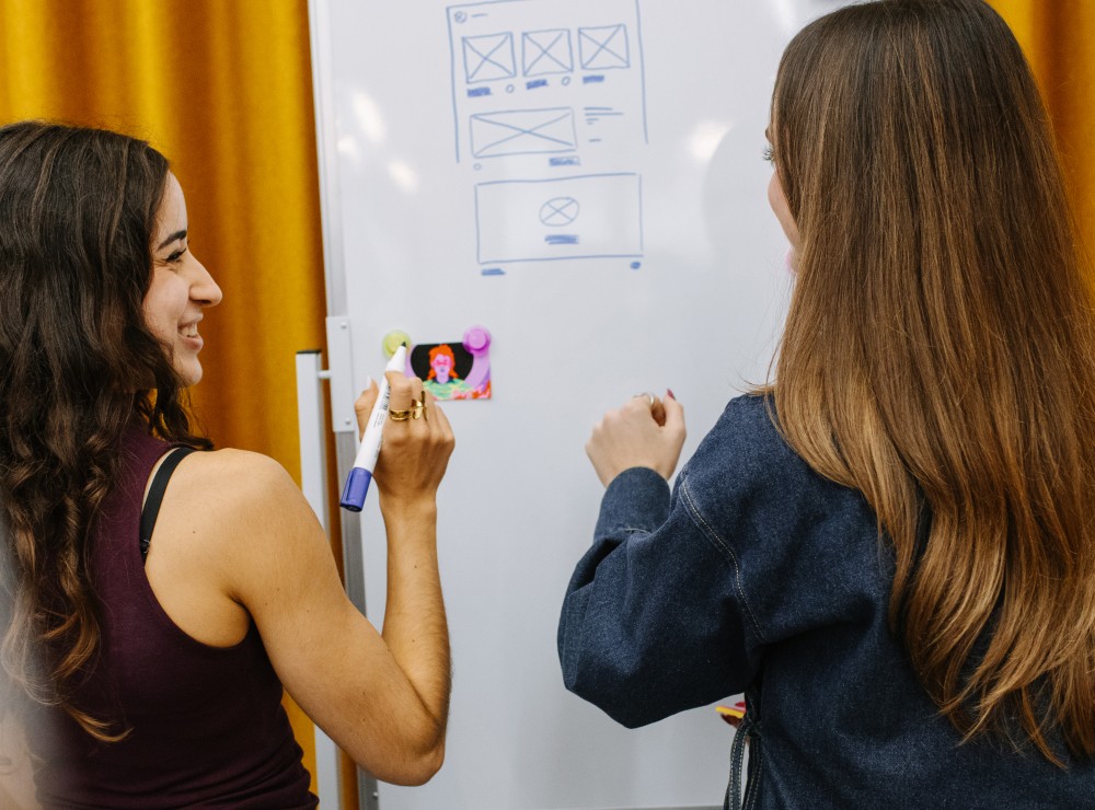 Two women reviewing wireframe sketches on a whiteboard in a workshop setting.