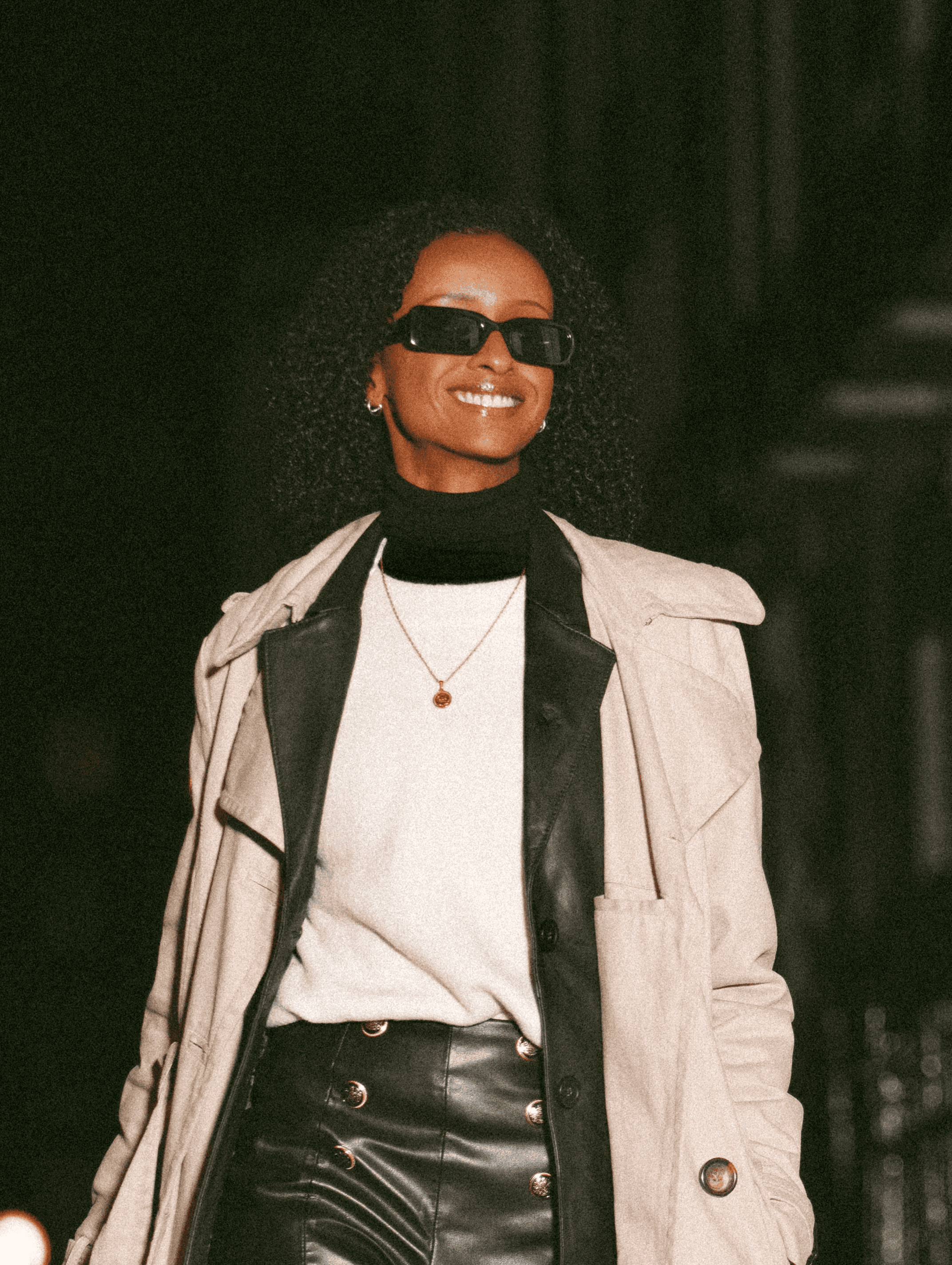 Smiling young woman with good-looking dark skin, wearing sunglasses and a fashionable outfit, standing outside at night.