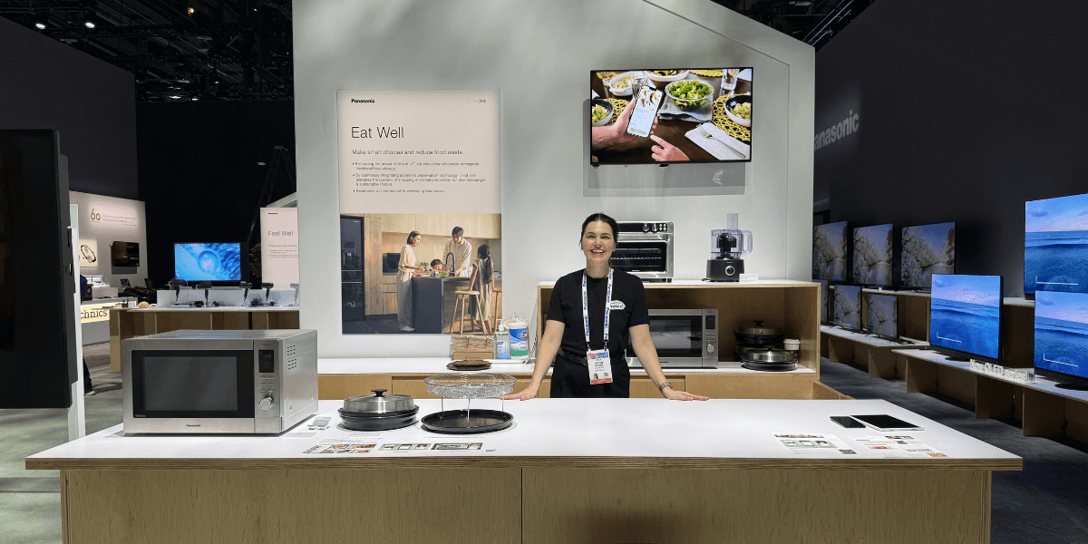 Fresco employee stands behind a Panasonic booth at a trade show with appliances, screens, and signage reading "Eat Well"