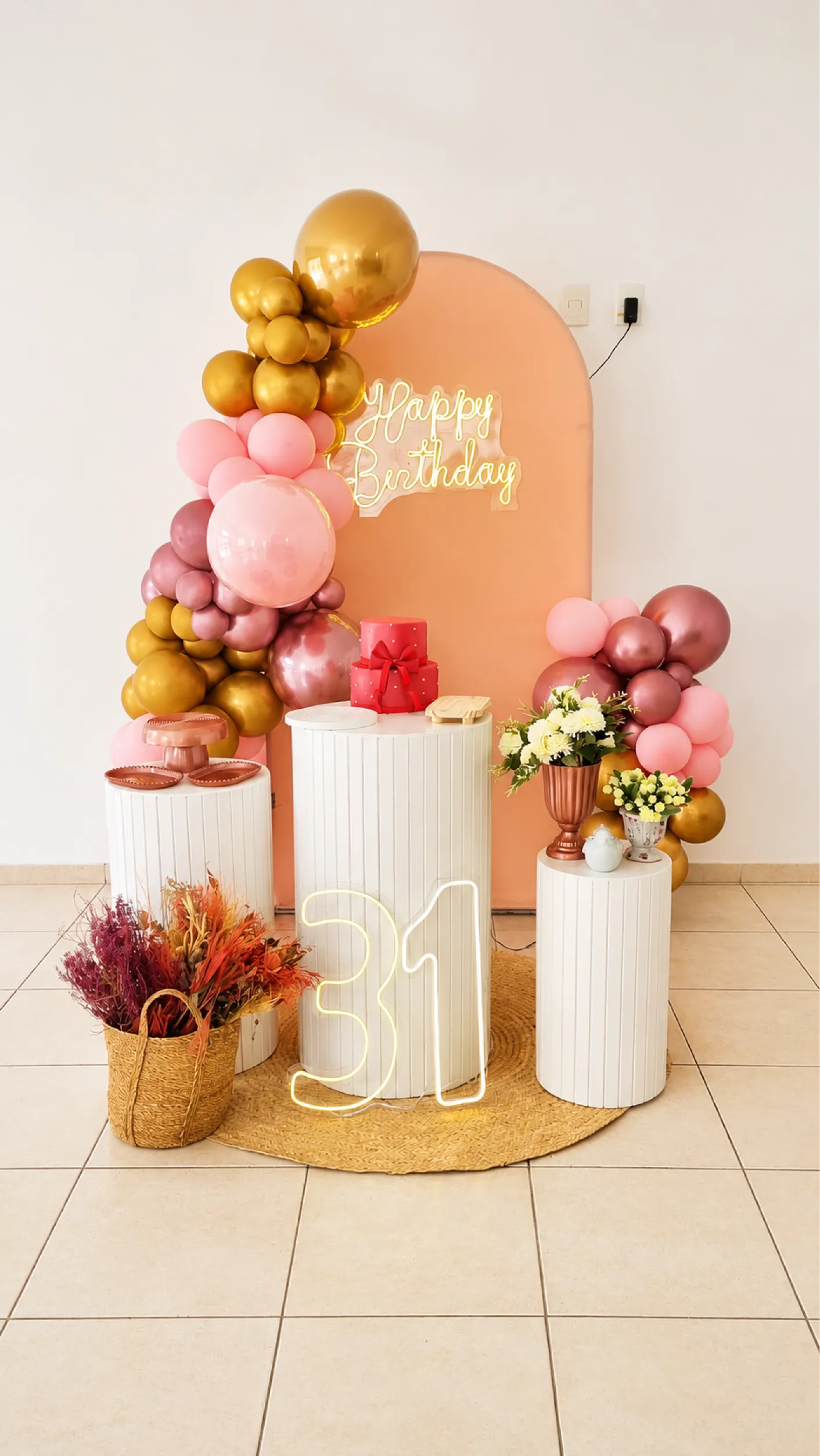 Decoração de aniversário em tons rosé, dourado e pêssego, com painel arredondado, balões, cilindros brancos, flores e número iluminado.