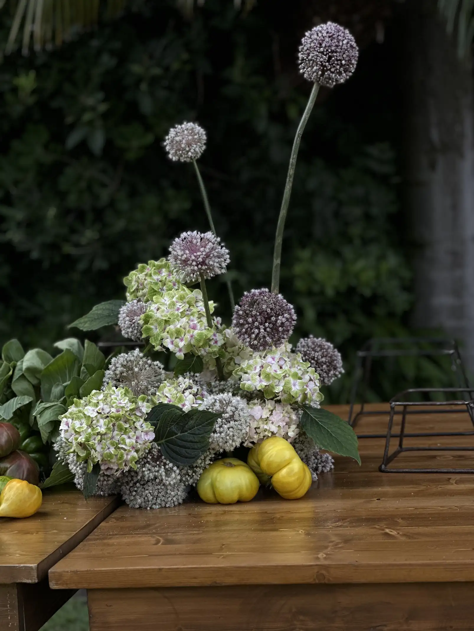 Arreglo floral con hortensias y flores esféricas lilas sobre mesa de madera para composición elegante de evento al aire libre.