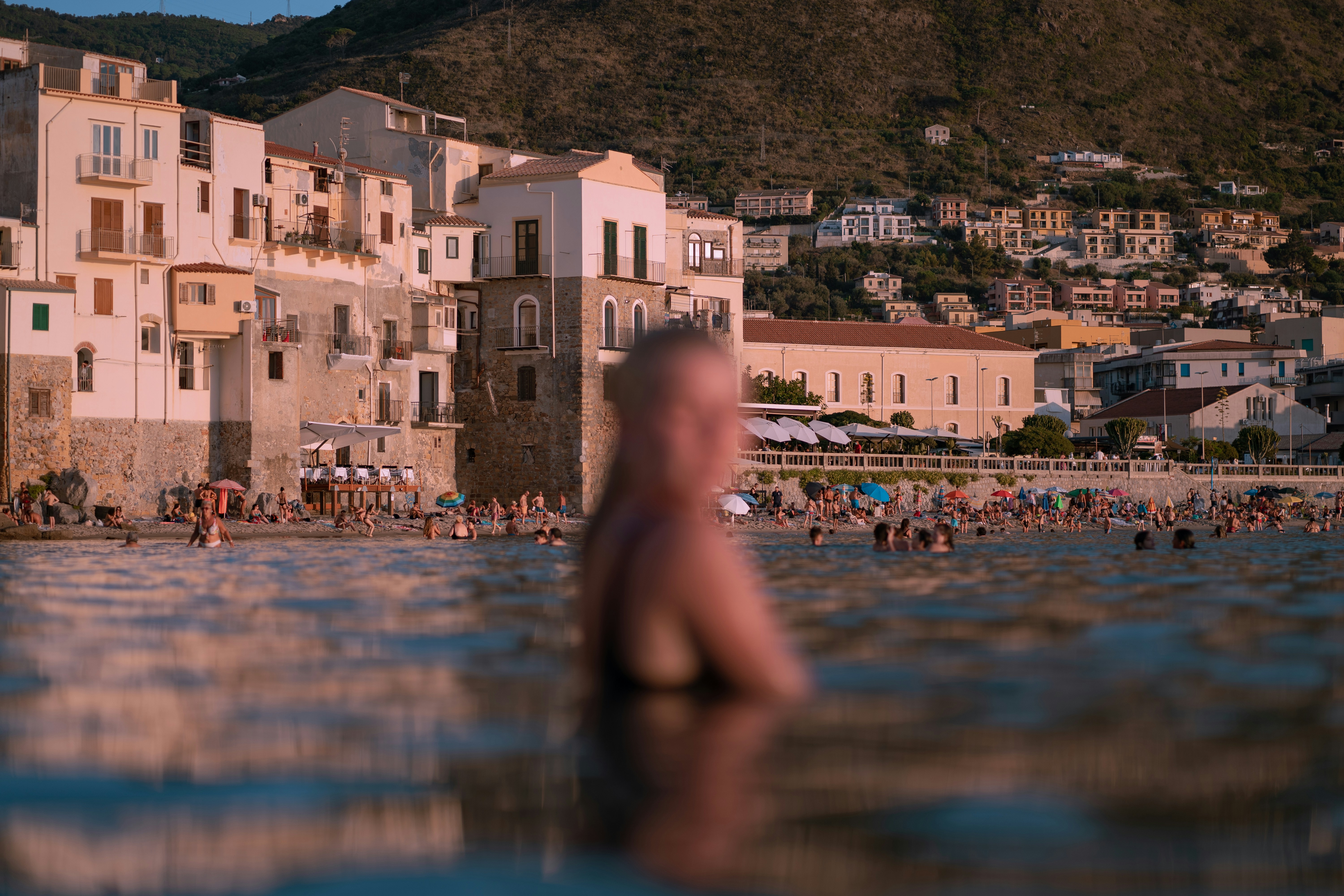 A person wades in water near a coastal village, with buildings and hills in the background during sunset.