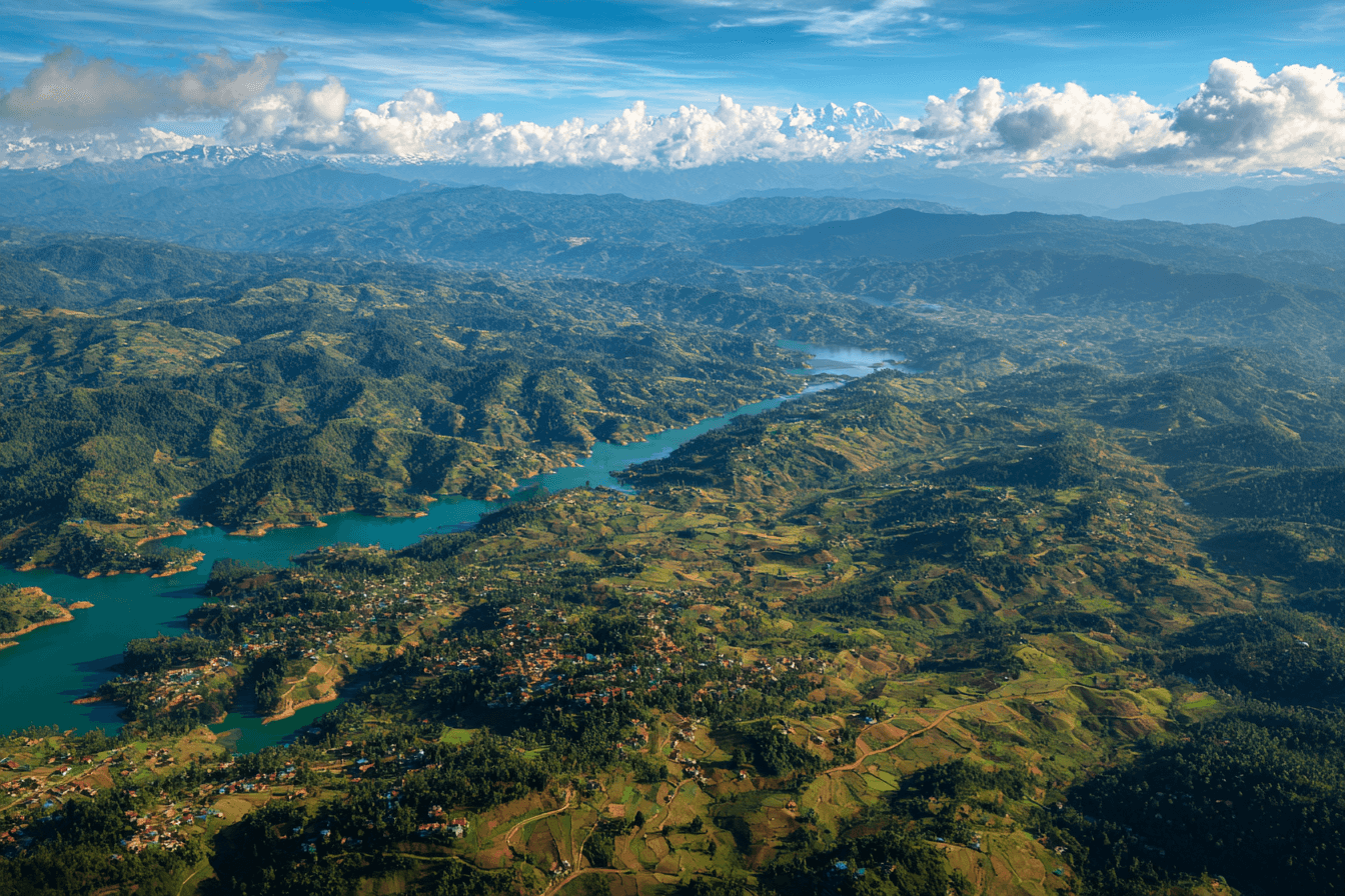 A breathtaking aerial view of a winding blue river cutting through lush green valleys and hilly landscapes, set against a backdrop of distant mountain ranges under a partly cloudy sky.