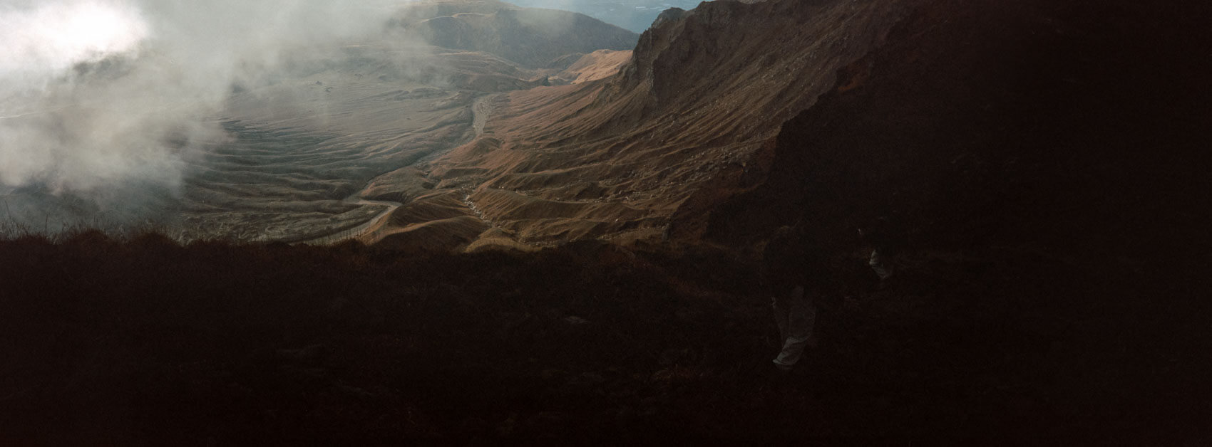 Japan hiking at Mount Aso: two hikers walking near an edge which leads to the volcanic crater, showing a high contrast environment, with humans for scale, captured on film in Kyushu's otherworldly terrain.