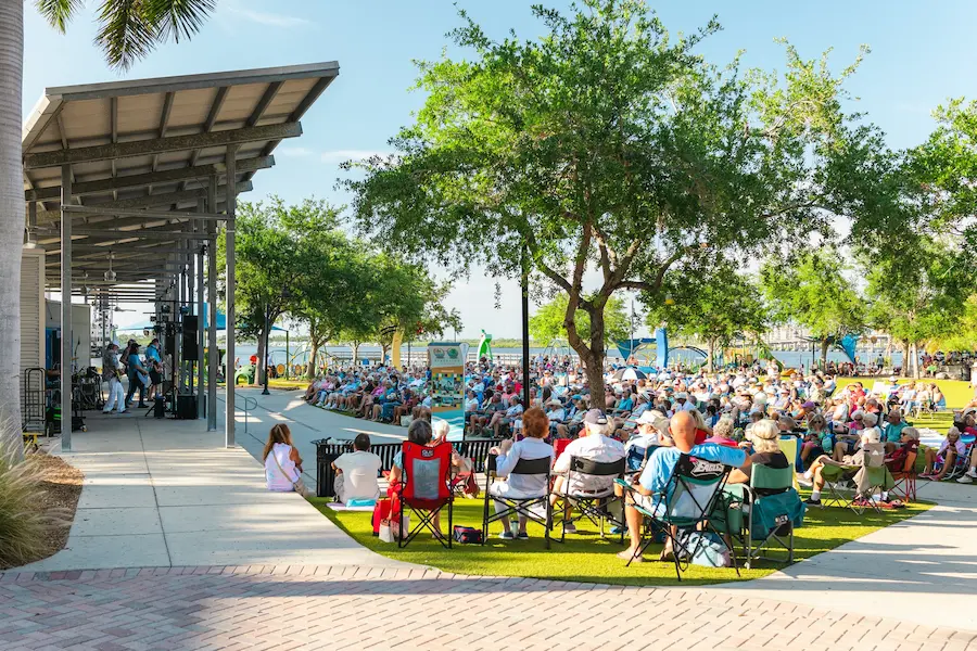 A large crowd gathers at an outdoor amphitheater in Bradenton, FL, surrounded by lush greenery and overlooking a serene waterfront, as they enjoy a live performance under a partly cloudy sky.