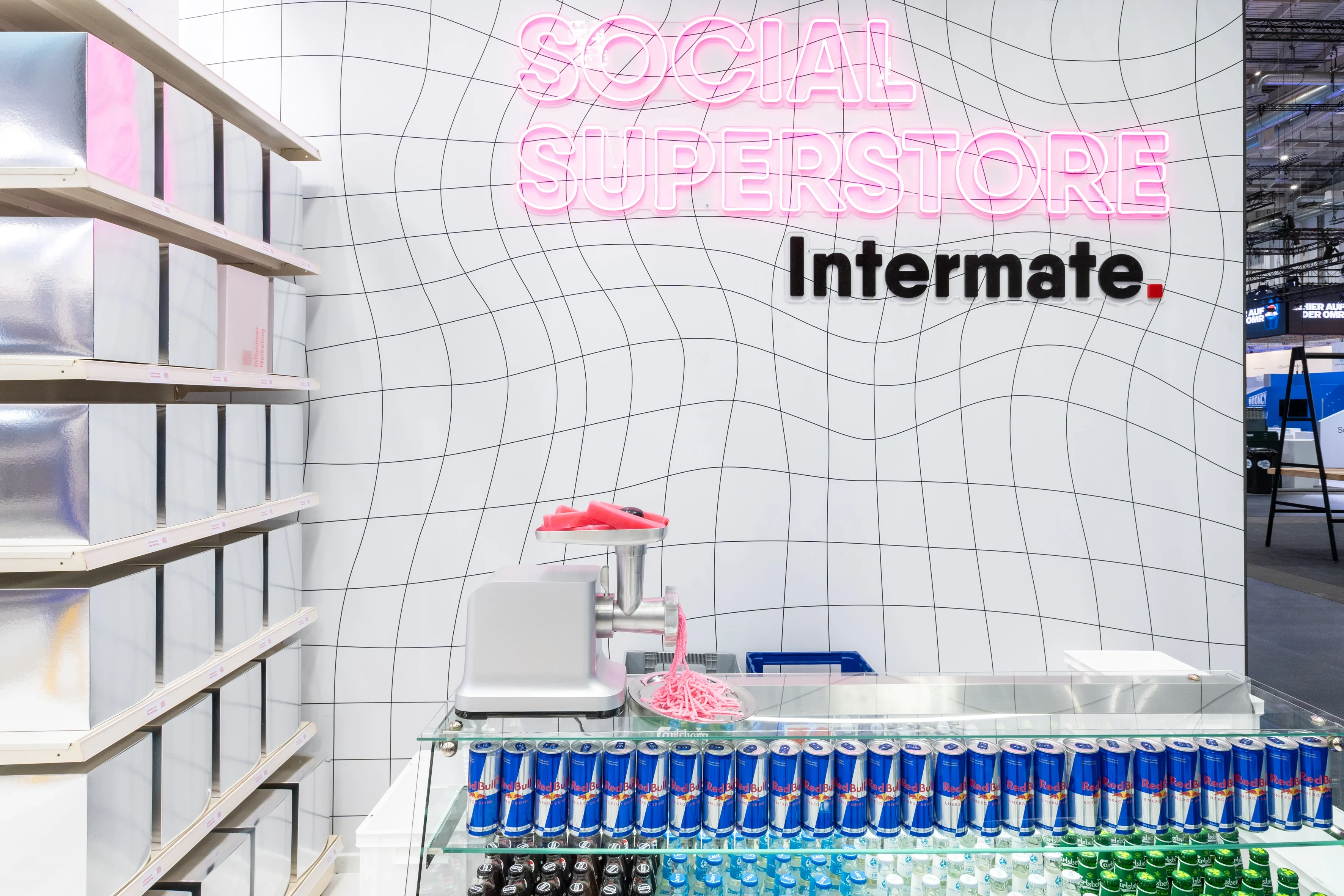 Meat grinder with pink content strands on a counter of Red Bull cans beneath the Social Superstore neon logo.