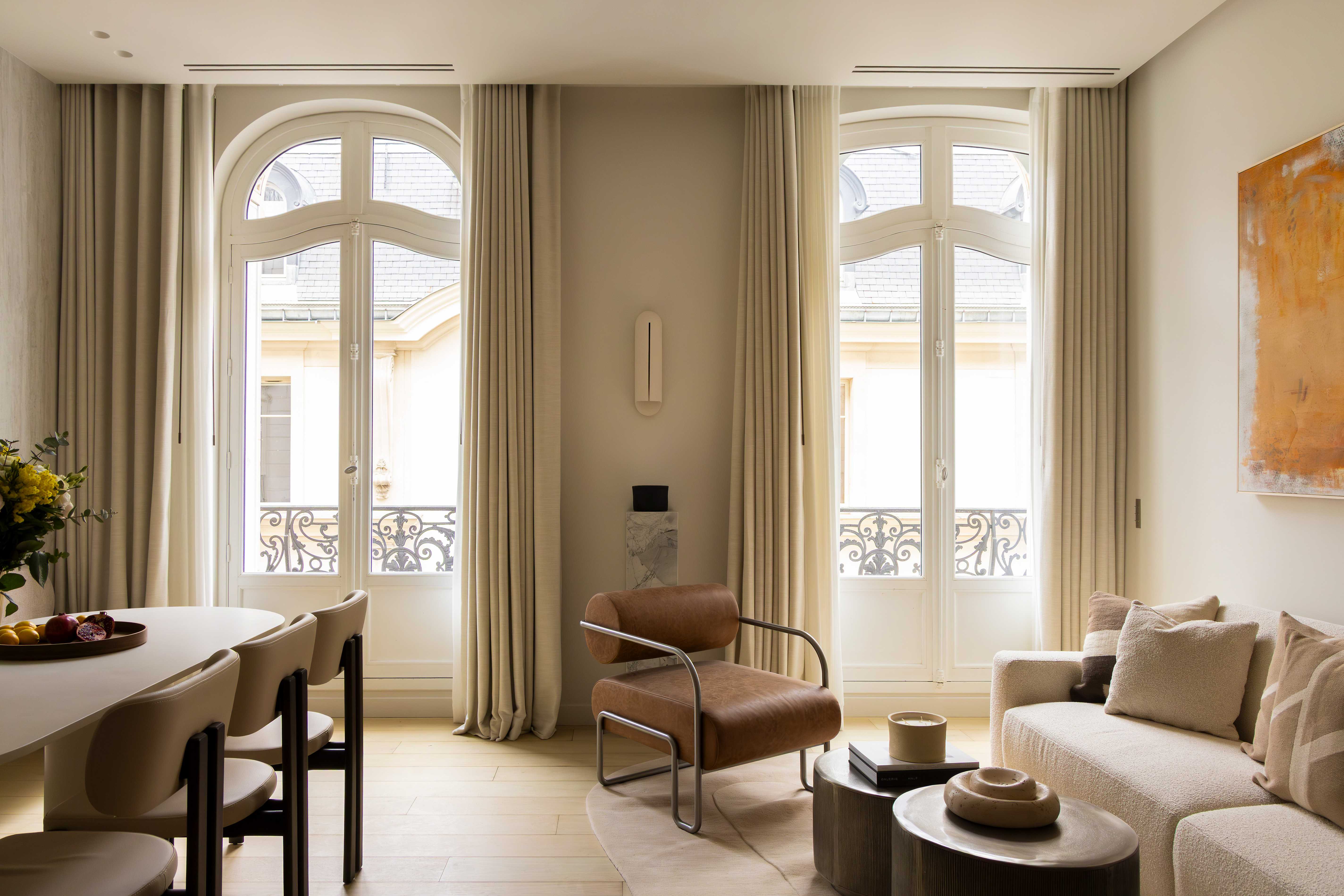Salon vu de face avec deux grandes fenetres cintrees, coin repas, fauteuil cognac et canape creme