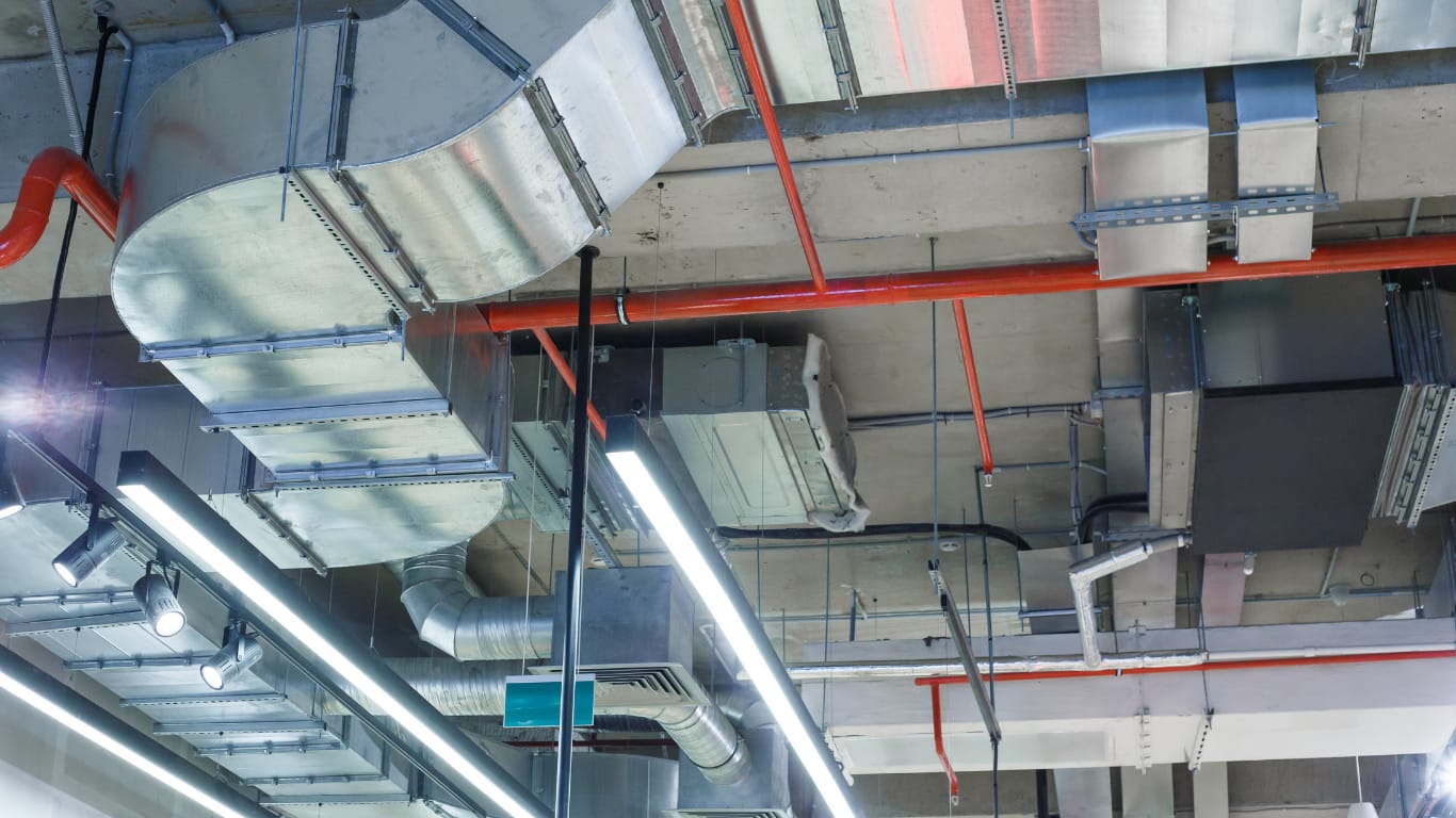 Blick an eine Industrie-Decke mit verschiedenen verzinkten Lüftungskanälen und einer auffällig rot lackierten Rohrleitung, die das System kreuzt. Lange LED-Lichtleisten erhellen die Installation.