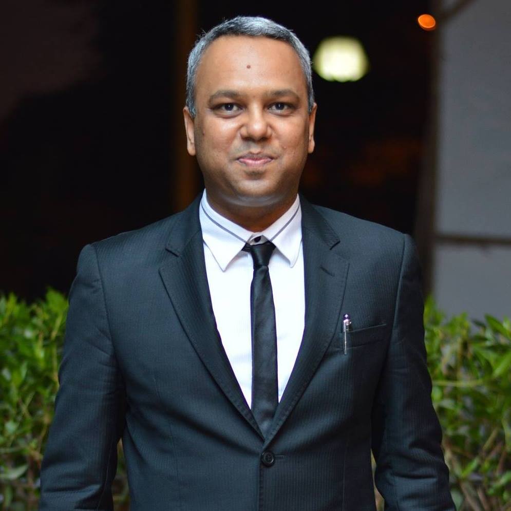 Smiling person with curly hair, wearing a black shirt, against a warm, dark background.