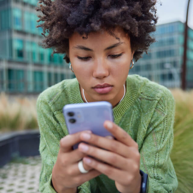 A person with curly hair wearing a green sweater sits outdoors, looking intently at a smartphone they are holding with both hands.