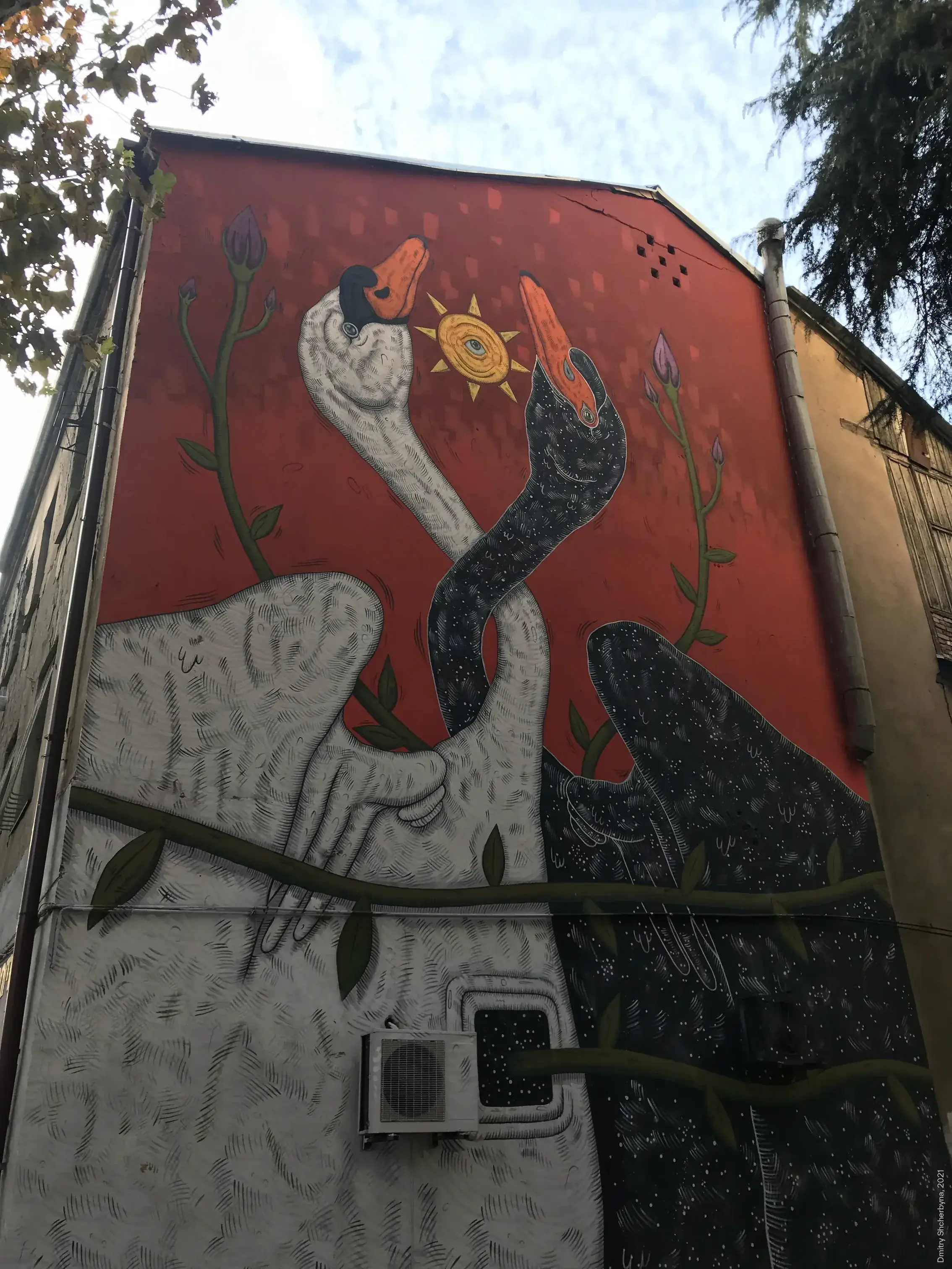 A mural on the side of a building showing two swans with intertwined necks — one white and one black speckled with stars — facing each other beneath a golden spiral sun against a deep red background.