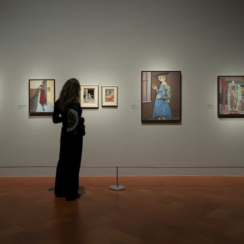 A person observing framed paintings on display in a dimly lit gallery with wooden flooring.