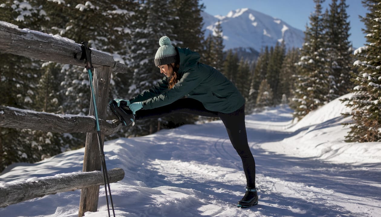 Skier stretching beside a snowy trail near a pine forest