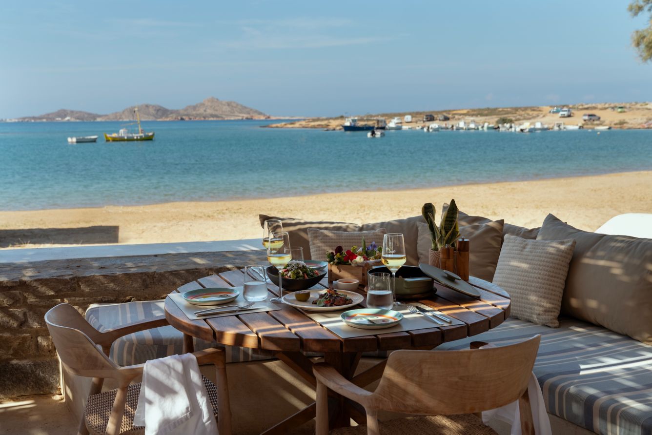 Laid round wooden table in the shade directly on a sandy beach with small fishing boats on the calm water.