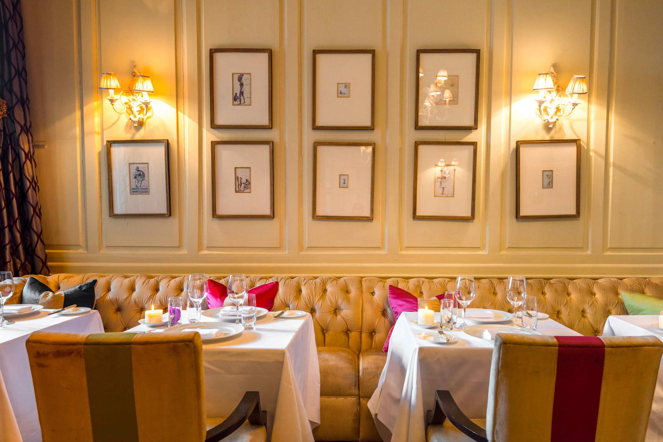 A row of tables with white cloths and colorful pillows in a restaurant with tufted seating and framed art on walls.