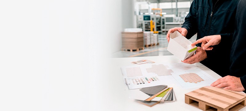 Progettazione del packaging in azienda: due persone esaminano il prototipo di un astuccio con le fustelle stampate, la mazzetta colori e i campioni di cartone, davanti a un pallet di scatole.