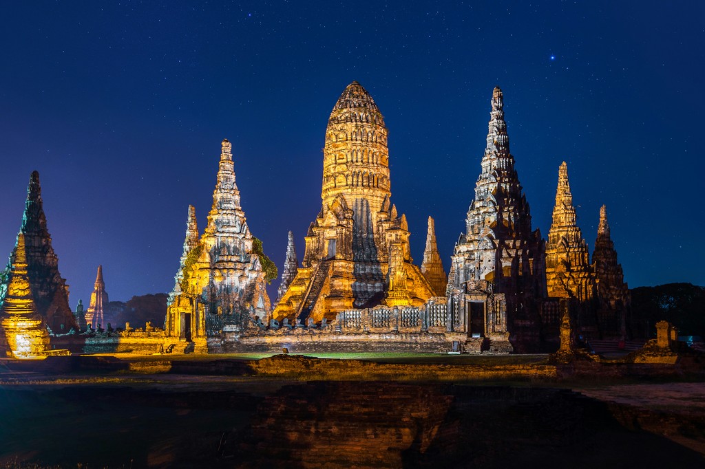 The illuminated ruins of Wat Chaiwatthanaram in Ayutthaya, Thailand, stand majestically under a starry night sky, showcasing the intricate architecture and historical grandeur of this ancient Buddhist temple complex.