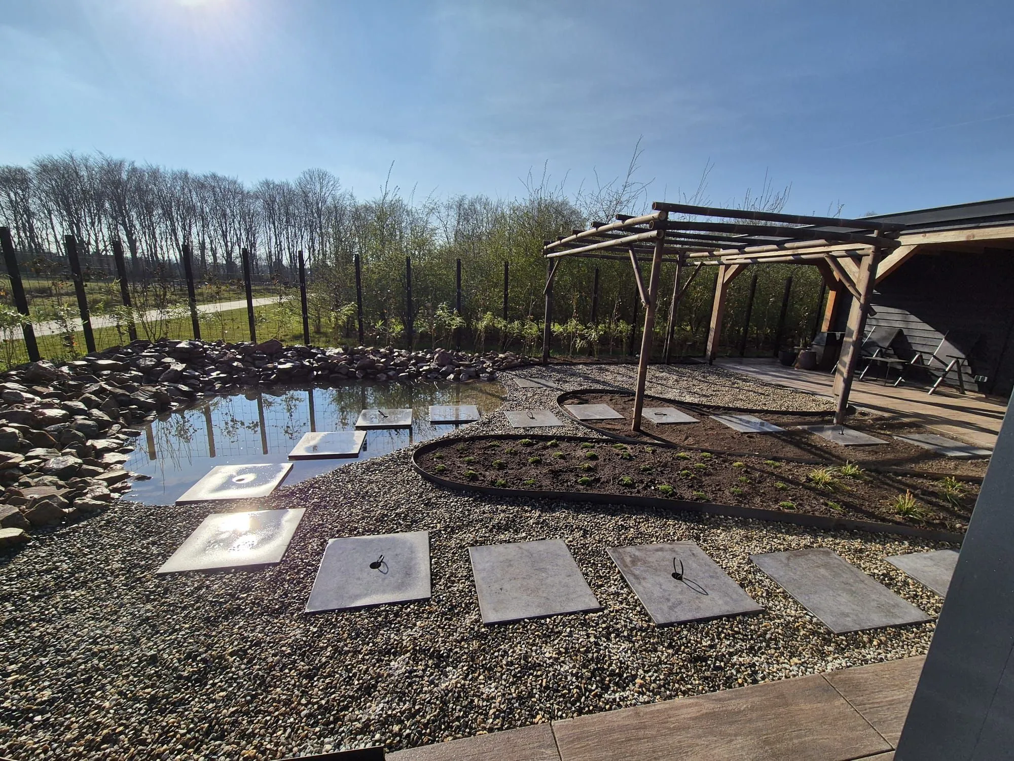 Rustige Japanse tuin met vijver omringd door rotsen, grindpad met grote betontegels en houten pergola bij nieuwbouwwoning in Gorredijk.