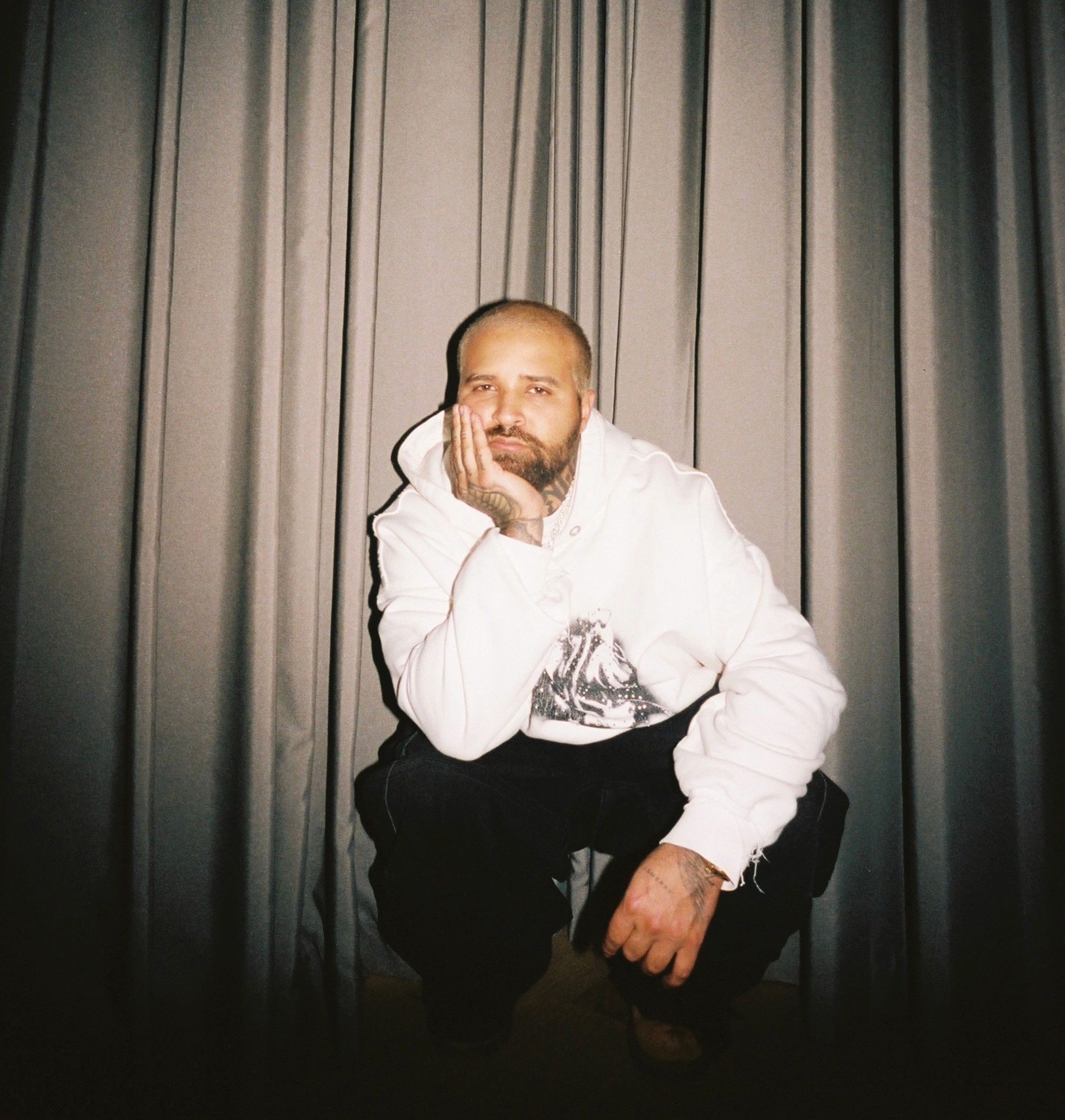 Azul Wynter, a music producer with a beard and tattoos, is seated in front of long gray curtains, wearing a white hoodie and dark pants, resting his head on his hand in a relaxed pose.