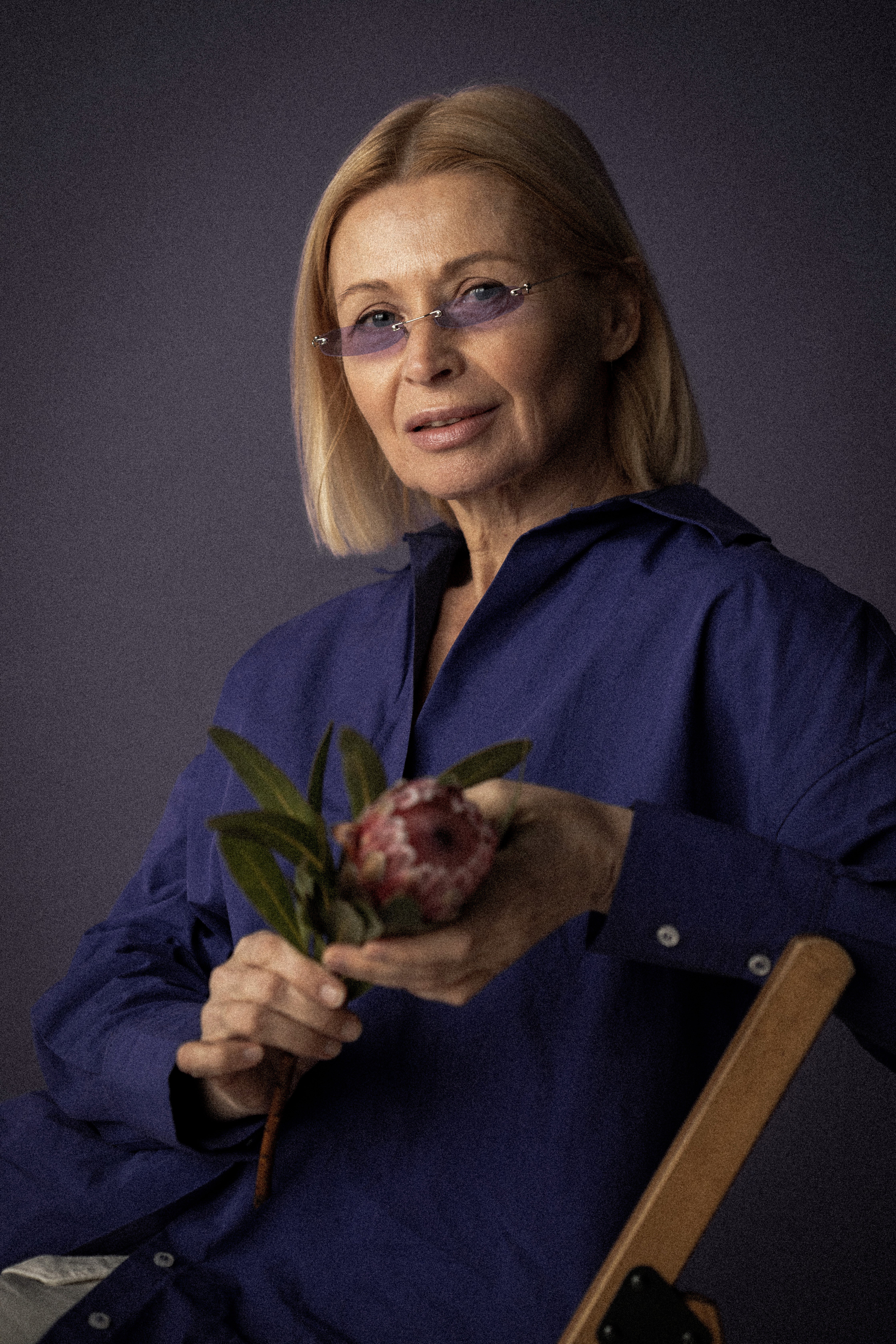 Une jolie femme d'âge moyen tenant une fleur et regardant la caméra, sur un fond violet foncé.