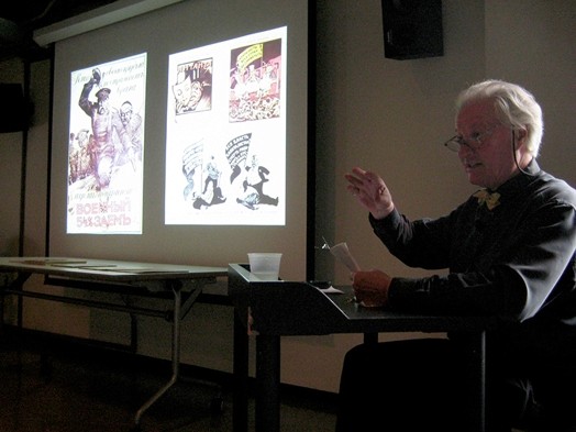 A photo of a man giving a lecture near a projection screen that shows what looks like old war propaganda.