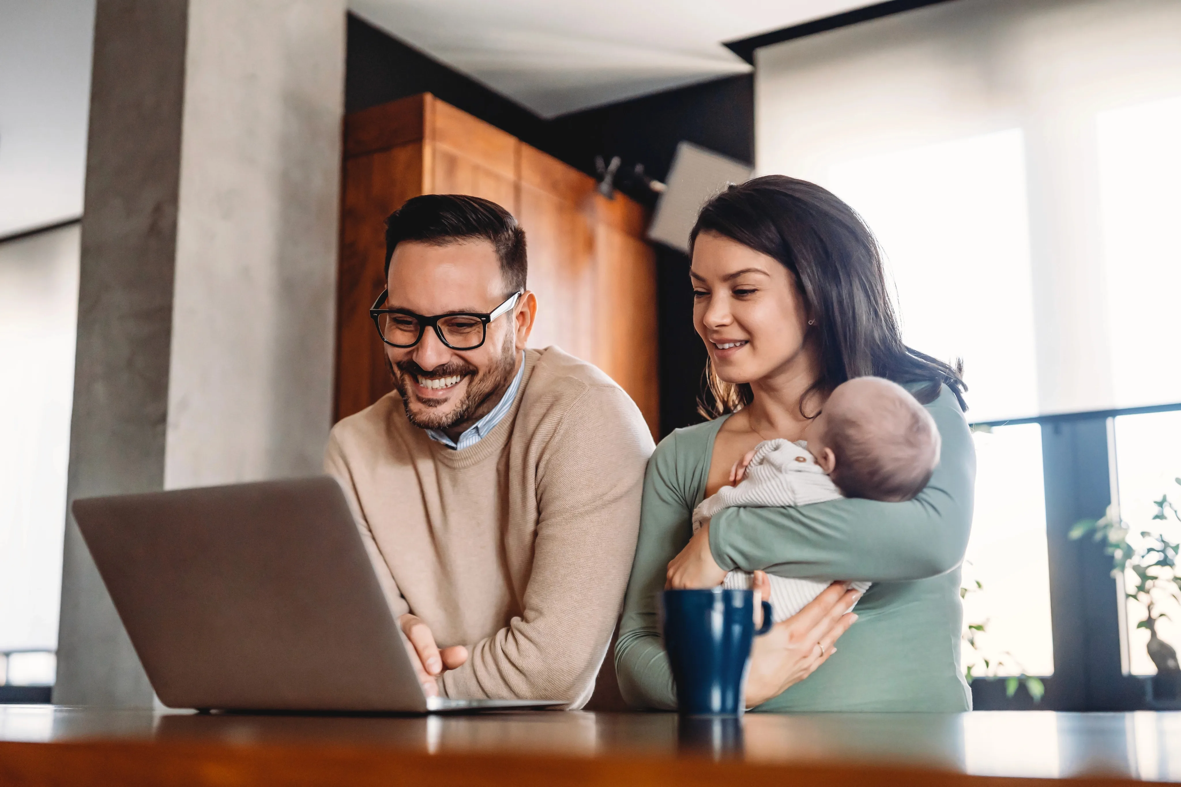 A young family smiling while looking at a laptop together, with the father wearing glasses and a beige sweater and the mother holding their newborn infant in a green top.