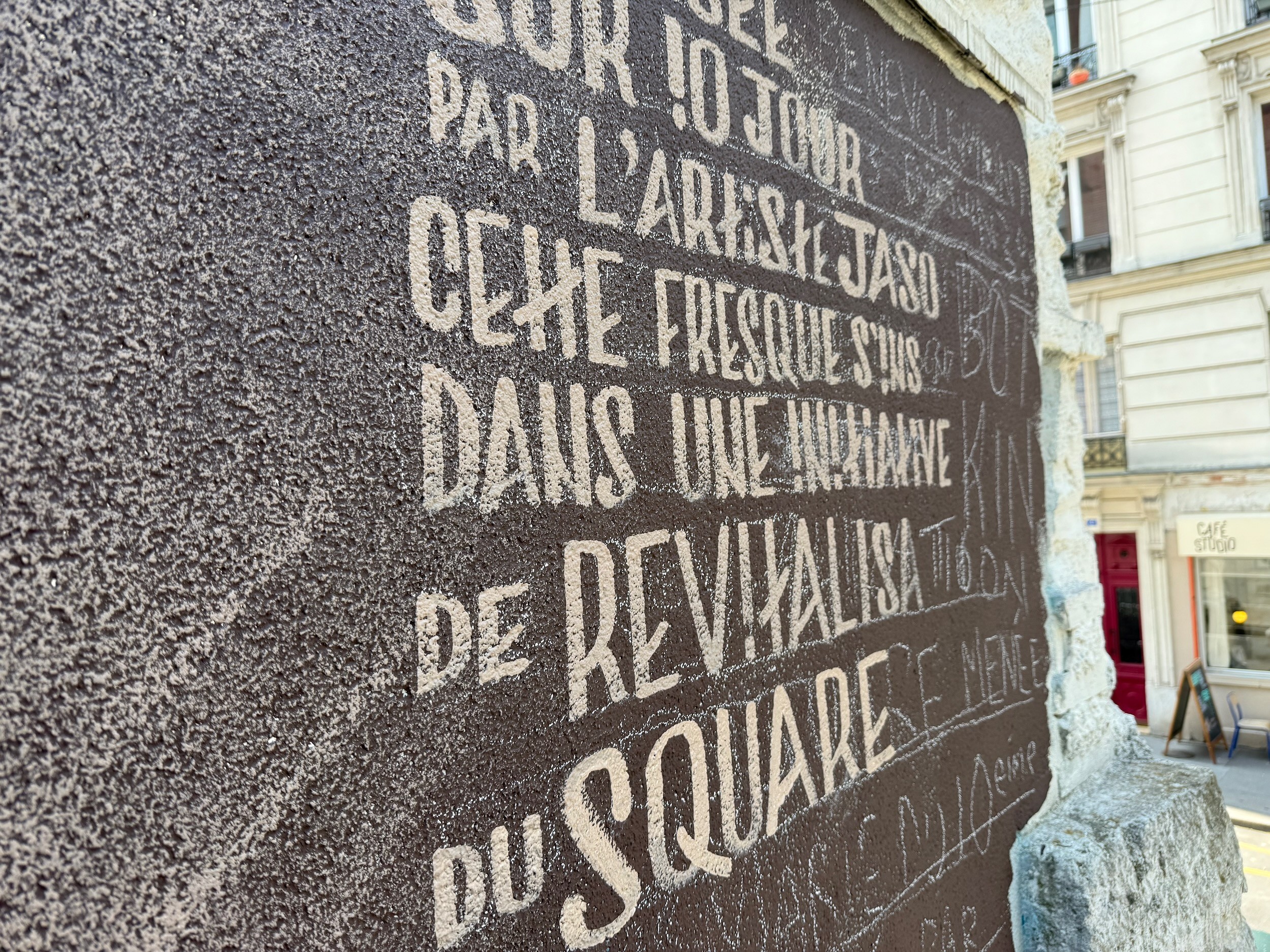 Close-up of white stencilled French lettering on the dark wall, describing the mural and the revitalisation of the square.