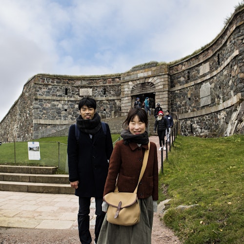 A man and woman are standing near the entrance of a stone fortress. Other people are walking up the path toward the structure.