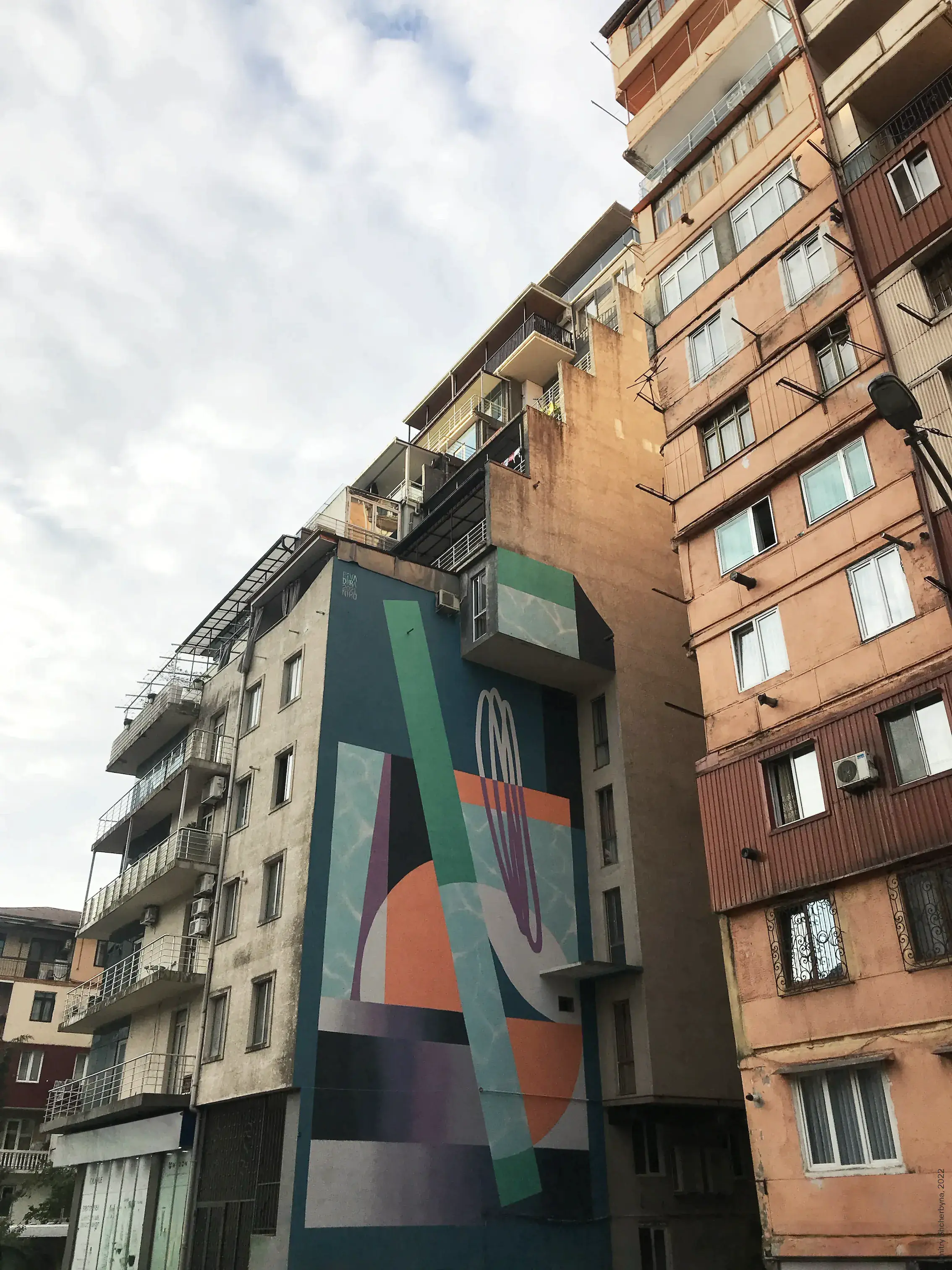 Abstract geometric mural painted in teal, orange, green and purple on the narrow side wall of a residential apartment block.