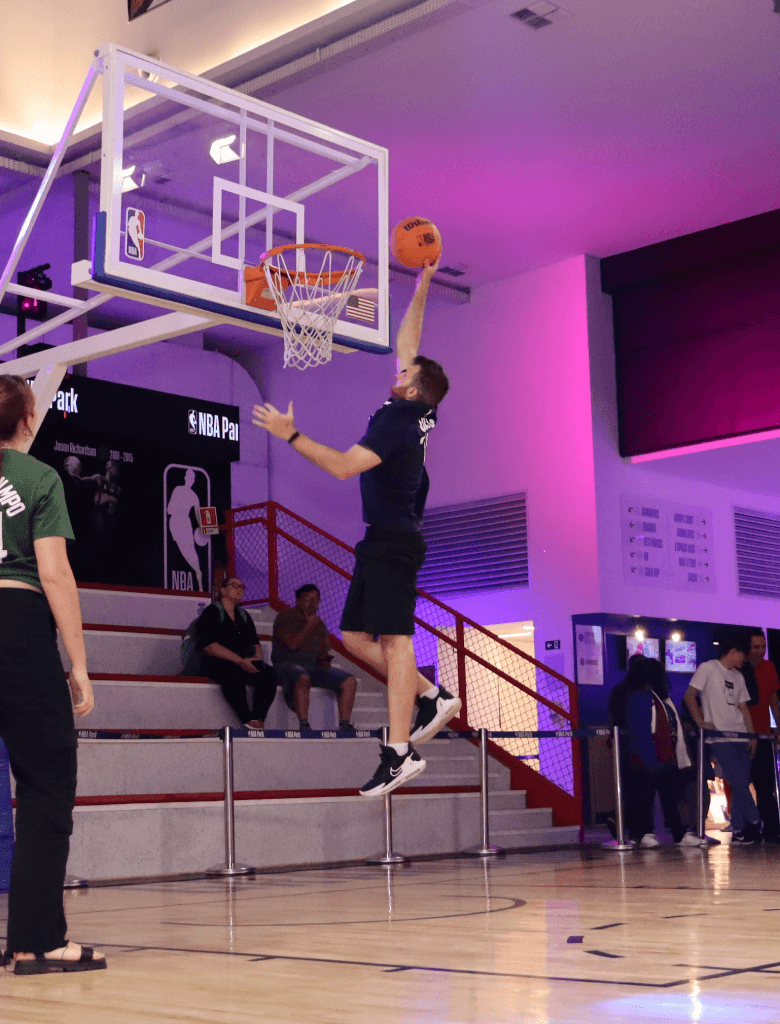 Augusto realizando uma enterrada em uma quadra de basquete interna, saltando em direção à cesta enquanto outras pessoas observam.